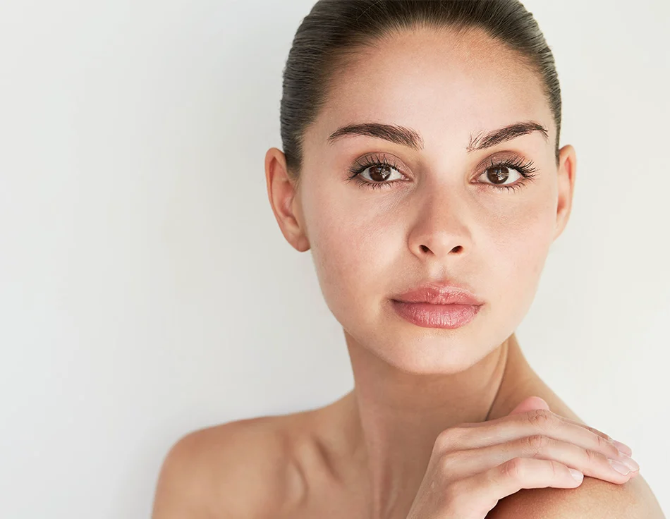 Close-up of a woman with minimal makeup, softly smiling with her hand gently resting on her shoulder, set against a light background - Exosomies in Houston, TX