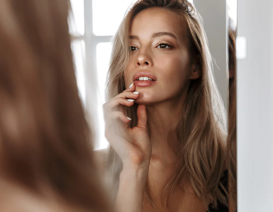 A woman with long blonde hair touches her lips softly, gazing at her reflection in a mirror, in a softly lit, intimate setting - Lip Lift in Houston, TX