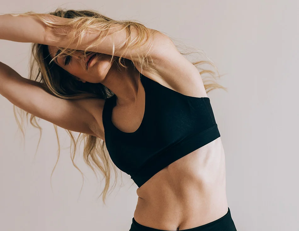 A woman with flowing blonde hair stretches with her arms above her head, wearing black sportswear in a minimalist, light-colored setting - Semaglutide in Houston, TX