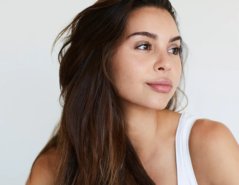 A woman with natural beauty and long, flowing hair looks off to the side with a relaxed expression, wearing a simple white tank top - Skin Pen Microneedling in Houston, TX