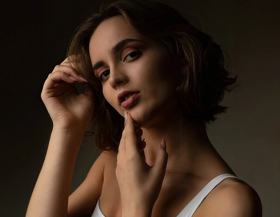 A woman with short, dark hair poses in low light, delicately touching her face with a serious, contemplative expression - Submental Liposuction in Houston, TX